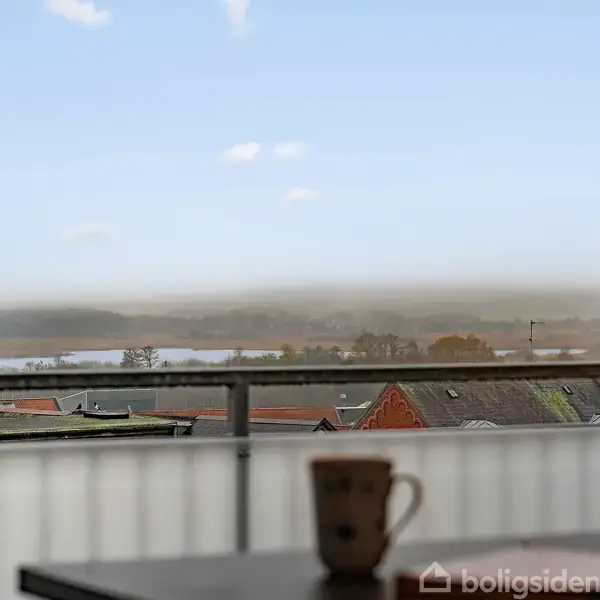 En kop står på et bord i forgrunden. Bagtæppet er en panoramaudsigt med tage og en sø i det fjerne, set gennem en altandag.