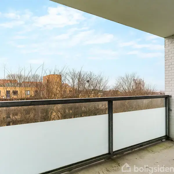 Balkon med glasrækværk rummer udsigt over vintertræer og omkringliggende bygninger under en blå himmel. Vægge af hvid mursten omkranser rummet.