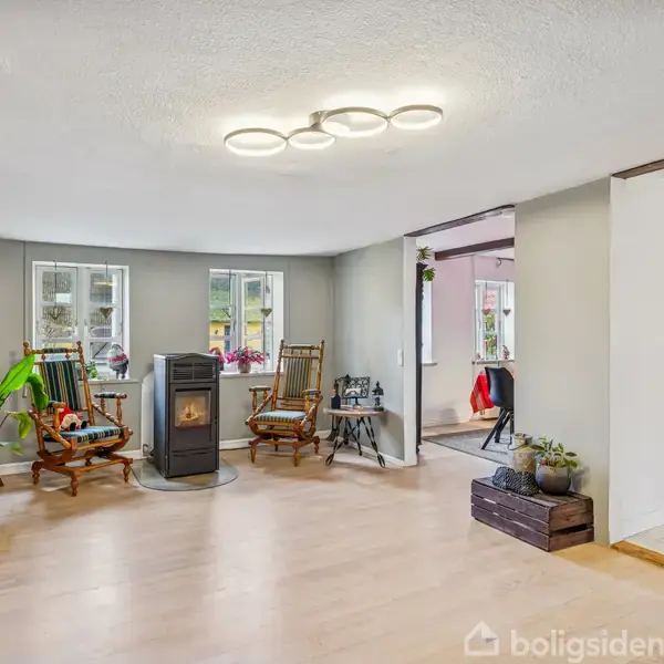 En stue med en brændeovn midt i rummet og to gyngestole ved siden af, omgivet af planter. En julemandfigur står i hjørnet. En åbning fører til et andet rum.
