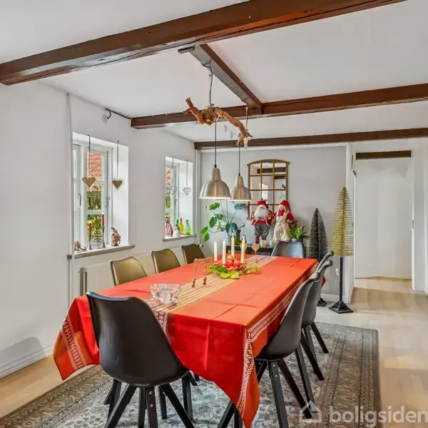 Langt bord dækket med rød dug, pyntet med stearinlys og dekorationer. Omgivet af sorte stole i en lys spisestue med bjælker i loftet og billeder på væggene.