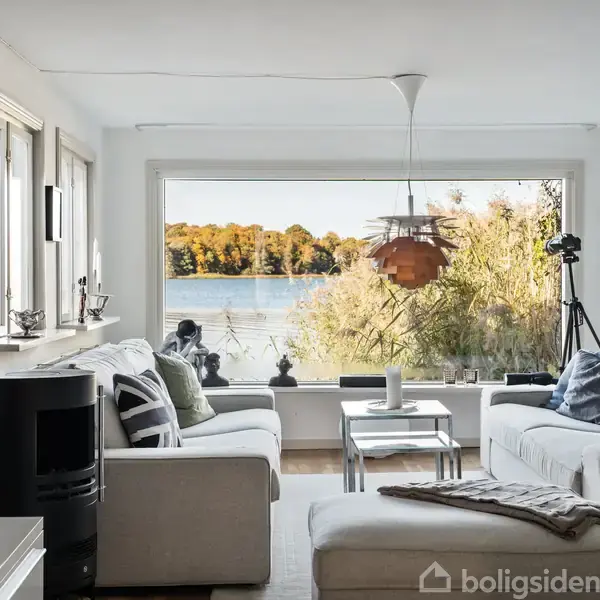 En stue med lyse sofaer og et sofabord står ved et stort vindue. Udenfor ses en sø med efterårstræer. Der er en kamin og en lampe indeni.