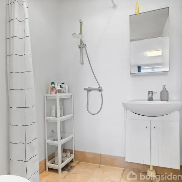 En hvidt badeværelse med en bruser til venstre, en håndvask med spejl over til højre, og et lille hyldemøbel med toiletartikler imellem. En bruseforhæng hænger ned ved bruseren.
