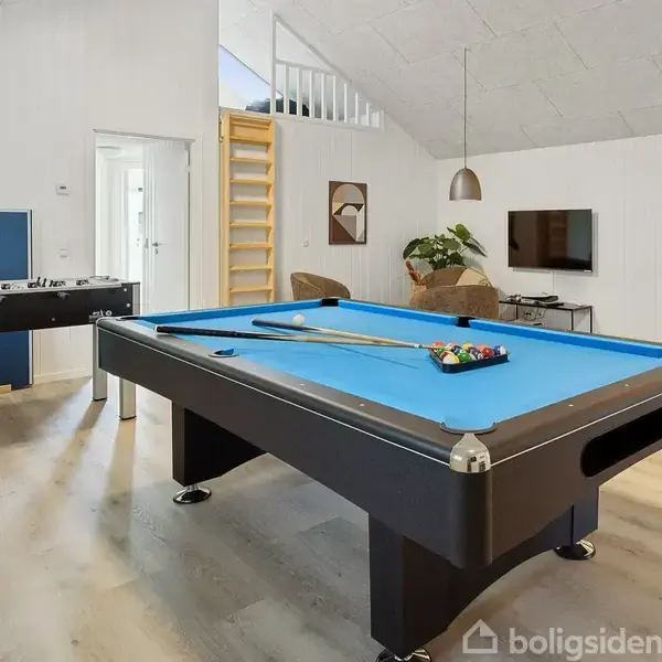 Poolbord står midt i et lyst rum med trægulv, omgivet af et bordfodboldspil, sofa, fladskærms-tv på væggen, og en reol. Loftet er skråt, og en lampe hænger ned.