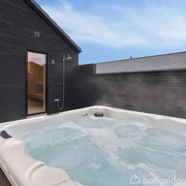 Udendørs jacuzzi bobler med damp i en moderne, afskærmet terrasse. En brusekabine er monteret på en mørkegrå murstensvæg. Himlen er klar og blå.