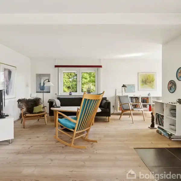 Stue med en gyngestol foran en pejs, der brænder. Omkring står sofaer, et skrivebord med lampe, malerier på væggene og et vindue med grøn udsigt.