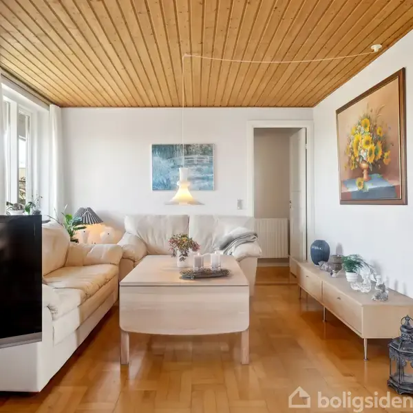 En stue med lys hjørnesofa omkring et sofabord, omgivet af malerier på væggene. Et tv står i forgrunden. Rummet har trægulv og træloft, med udsigt til en gang.