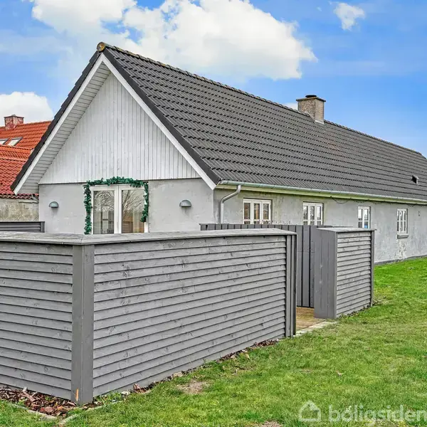 En lang, hvidmalet hus med mørkt tag står omgivet af en grøn græsplæne. Foran huset er en grå, træhegn indhegning. Himlen er blå med få skyer.