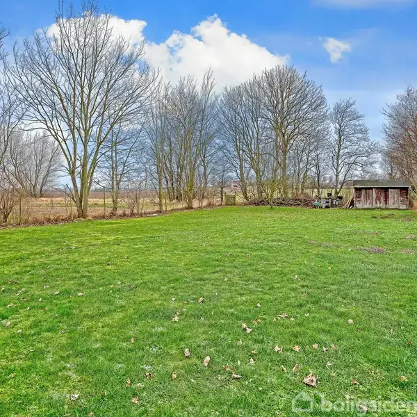 Græsplæne omgivet af nøgne træer, strækker sig mod en lille træbygning i baggrunden. Himlen er blå med enkelte skyer, og omgivelserne virker landlige og rolige.