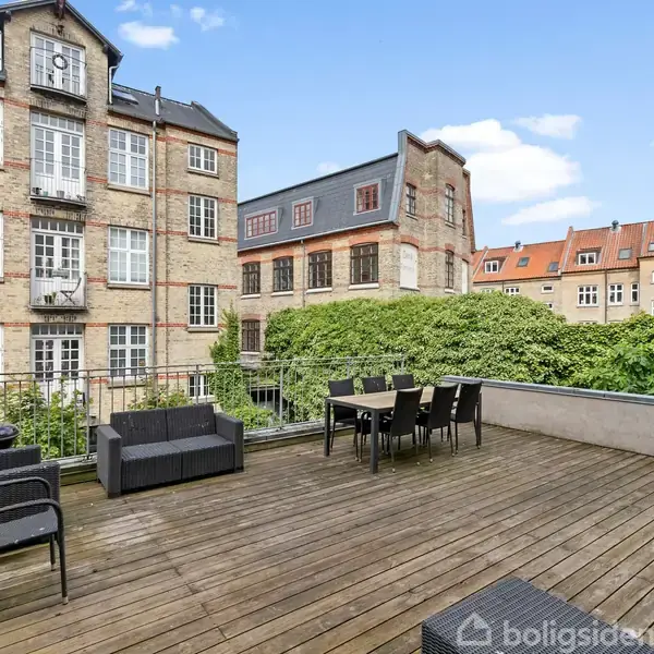 Stor, træbelagt tagterrasse med sort udendørsmøblement, inklusive stole og et bord. Omgivet af murstensbygninger med mange vinduer, under en skyfri blå himmel.