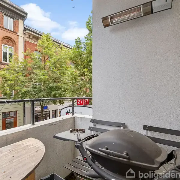 Grill på lille altan; står stille ved et bord under en varmeblæser. Omgivet af bymiljø med murstensbygning, træer og en gadeskilt markeret "27/21".
