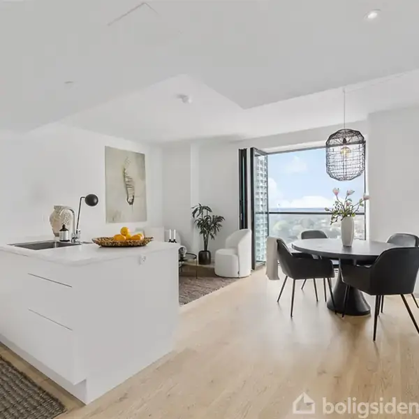 Et moderne, lyst køkken-alrum med et hvidt køkkenø bord med en frugtskål. I baggrunden er der et rundt spisebord med sorte stole og en glasdør til balkon.