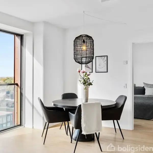Et rundt spisebord er omgivet af fire sorte stole og dekoreret med en vase med blomster, stående i et lyst rum med store vinduer, under en loftslampe af flet.