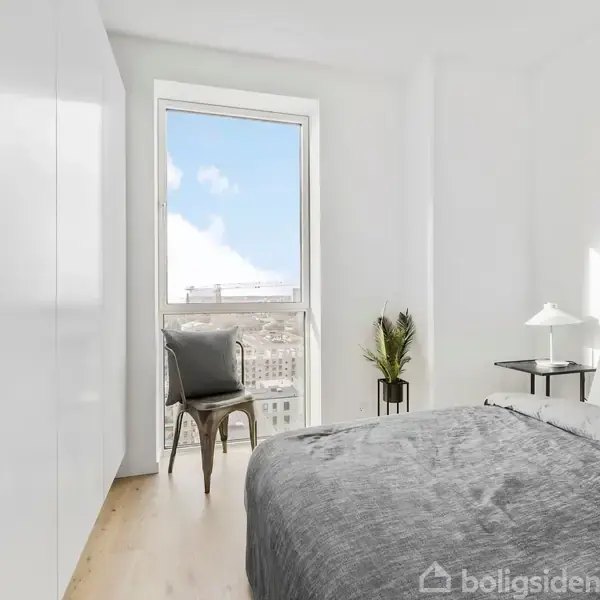 Et minimalistisk soveværelse med en grå seng til højre, en stol ved et vindue i midten, og en hvid garderobe til venstre. Indretningen er lys og moderne.