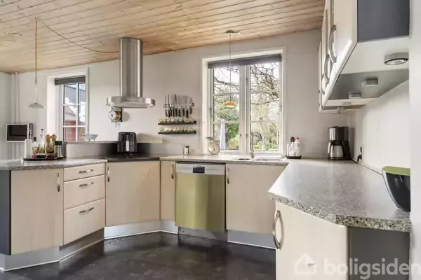 Et moderne køkken med lyst træloft. Køkkenelementer er lyse med en indbygget sølvfarvet opvaskemaskine. Vinduerne giver udsigt til en grøn have. Rummet har mørkt gulv og rustfrit stål belysning.