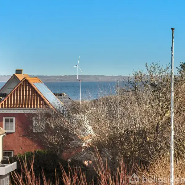 To huse står foran træer med udsigt til havet, hvor en vindmølle ses i det fjerne under en blå himmel.