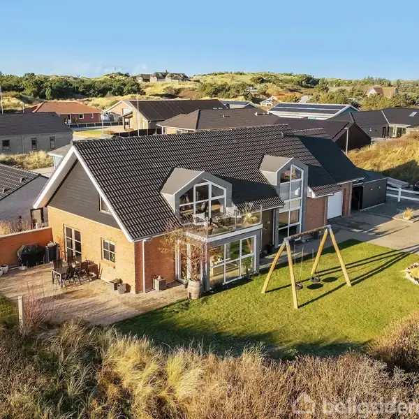 Hus med mørkt tag og røde murstensvægge, står stille på grøn græsplæne med gyngestativ foran. Omgivet af andre huse og sparsomt beplantet natur.