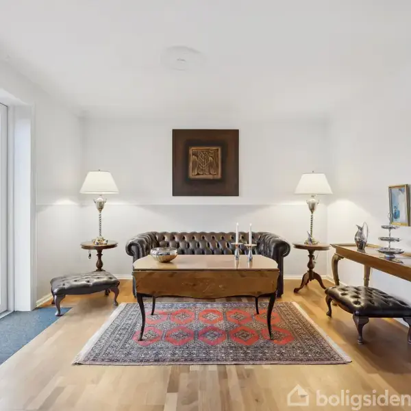 Et mørkt lædermøblement står i midten, flankeret af to lamper på små borde. Stuen har træmøbler, et farverigt tæppe og udsigt til en have gennem glasdøre.