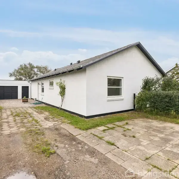 Et hvidt enfamiliehus har en flisebelagt indkørsel. Huset har en sort tagryg og en garage i baggrunden. Omgivet af græsplæner og buske under en klar blå himmel.