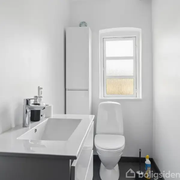 Hvidt badeværelse med håndvask og toilet. Håndvasken har spejl og sæbedispenser, og ved siden af er et højskab. Vinduet i midten giver lys til rummet.