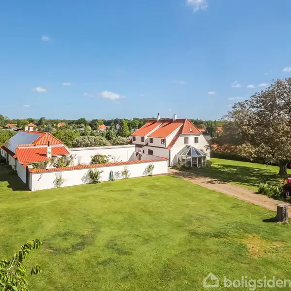 Et hvidt hus med rødt tag står omgivet af grønne marker og træer. En grusvej fører op til huset, der har en stor have med blomster og buske.