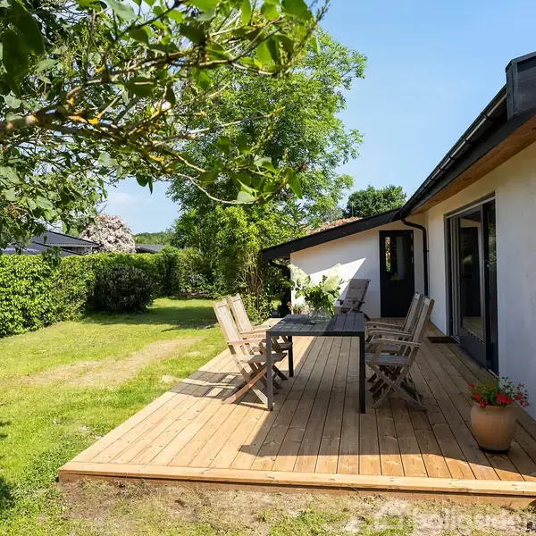 Terrasse med træbord og seks stole står på en træplatform. Omgivet af frodig have med græs og buskads under blå himmel. Peberplanter i krukke ved siden af huset.