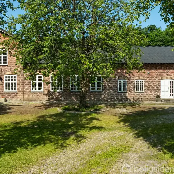 En stor, rektangulær murstensbygning med mange hvide vinduer og to døre. Et træ står centralt i forgrunden, med grønt græs og skygger omkring.