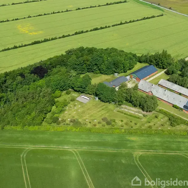 En gård med flere bygninger omgivet af marker og skov i et landligt område. Veje løber langs markerne, og en dam er synlig nær bygningerne.