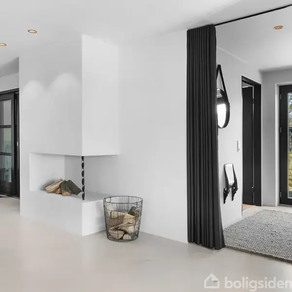 En stue med en hvid pejs og brænde i en kurv. En sort vindeltrappe leder til en etage, og store glasdøre fører ud til en terrasse med udsigt til natur.
