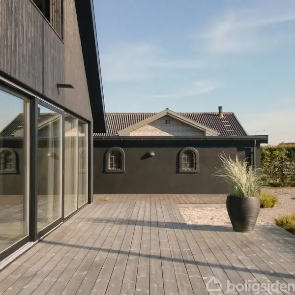 Moderne træhus med store vinduer åbner ud til en rummelig træterrasse. En stor urtepotte med grønt græs står på terrassen. I baggrunden ses en anden bygning og grønne hække.