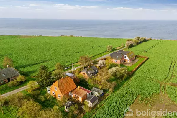 Gårdene ligger spredt i et frodigt, grønt landskab ved kysten, omringet af marker og tæt ved havet under en blå himmel.
