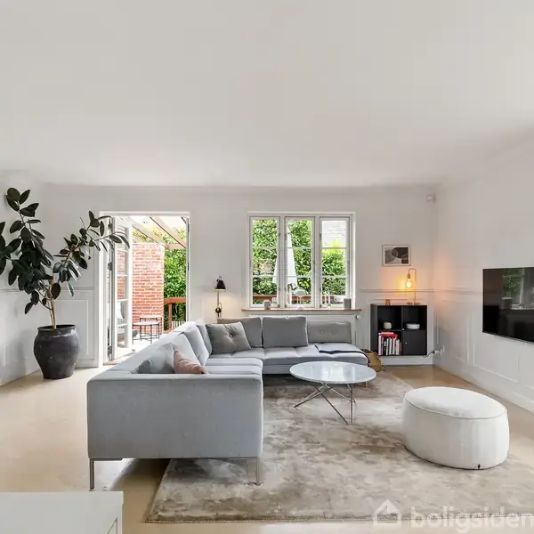 Grå sofa står i en lys stue ved et stort vindue. Fjernsynet hænger på væggen, og en stor plante i en krukke står ved siden af. En terrasse kan ses udenfor.