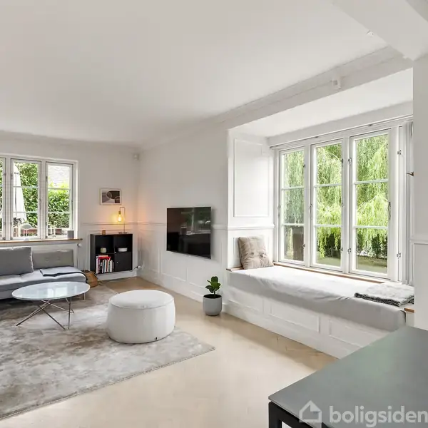 Stue med grå sofa og rundt sofabord, roligt oplyst med flere vinduer. Vindueskarmen har puder og et tæppe, og der står en lille grøn plante ved siden af. En TV-skærm hænger på væggen.
