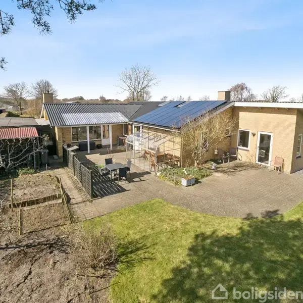 Hus med solpaneler på taget står i en velplejet have med græsplæne. En terrasse forbinder bygningerne, omgivet af buske og træer.