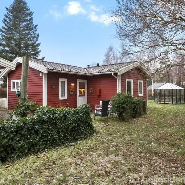 Rødt træhus med hvide vinduer og døre står stille i en have med en grøn græsplæne. Baghaven har et lille drivhus og omkranses af træer.