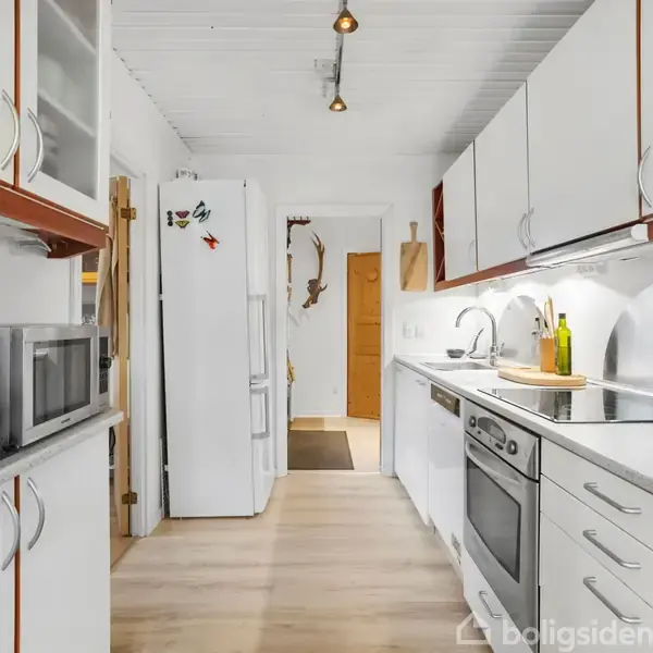 Et moderne køkken med hvide skabe og apparater, herunder en ovn og mikrobølgeovn. Køkkenborde i lyst træ. I baggrunden står et åbent køleskab, og lys skinner fra loftet.