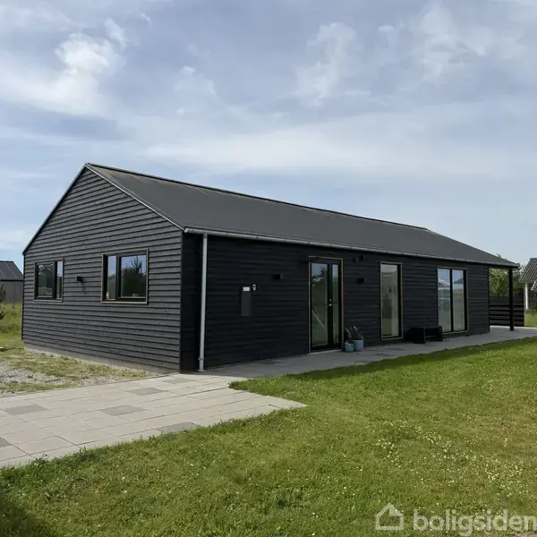 Et sort træhus står stille på en grøn græsplæne med fliser foran døren. I baggrunden ses flere huse under en blå himmel med lette skyer.