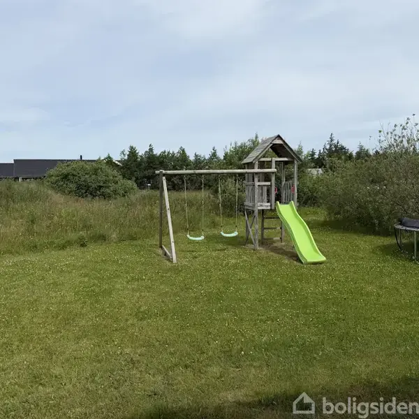 Et gyngestativ med to gynger og en rutsjebane står på en grøn græsplæne omgivet af buske og træer. En trampolin ses til højre i billedet.