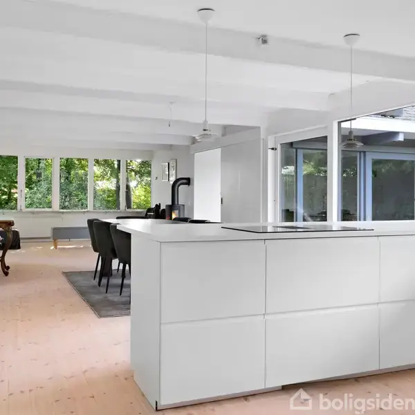 Et lyst køkken med hvidt skabsmodul, to hængelamper og store vinduer i baggrunden. Spisebord og stole ses ved siden af pejsen, med grønne træer udenfor.