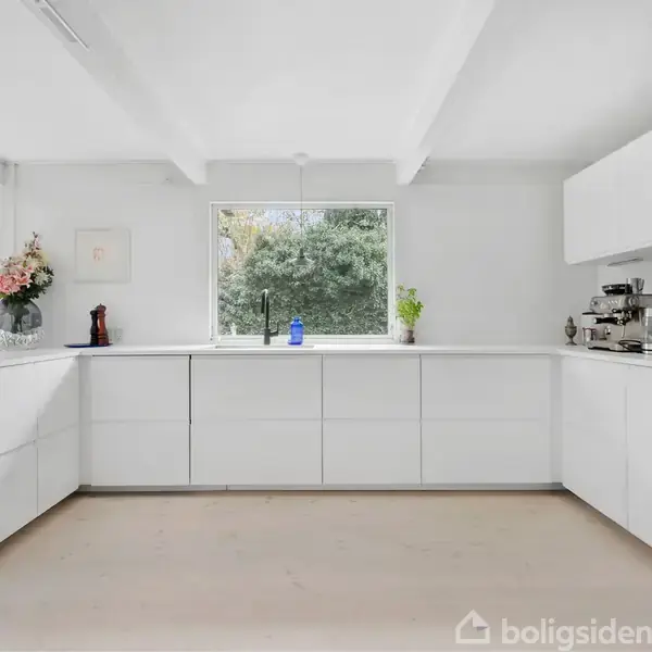 Hus ligger stille blandt frodige træer og blomstrende buske med lilla blomster. Skovklædt baggrund tilføjer en fredfyldt atmosfære.