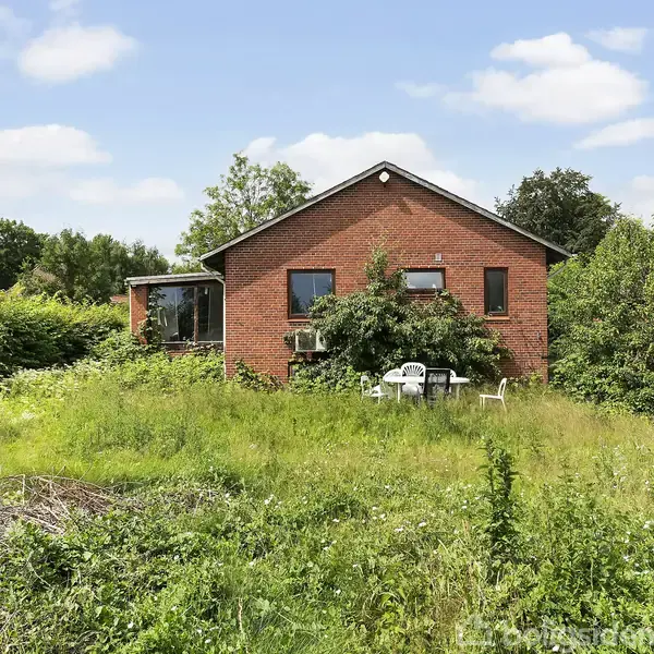 Rødt murstenshus har en tilgroet have med græs og buske. To hvide havestole står foran huset. Omgivet af andre huse og træer under en blå himmel med få skyer.