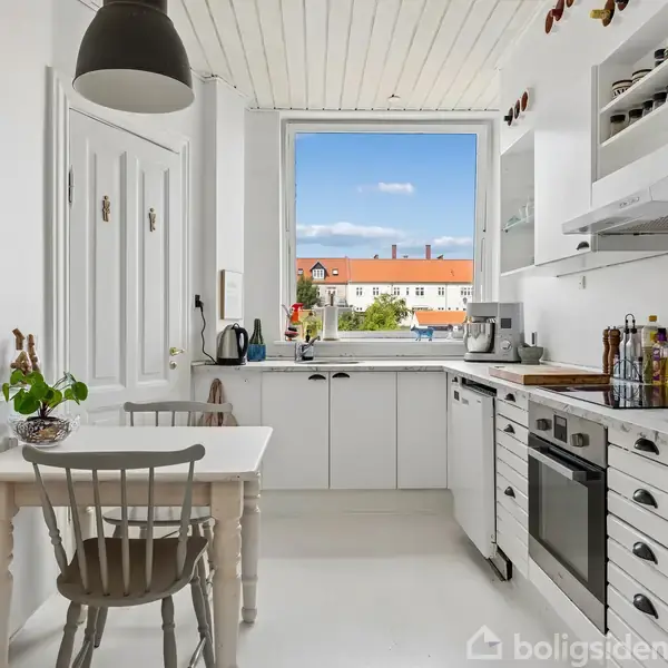 En gang med en hvid dør for enden. En bænk står langs væggen, mens frakker hænger på knager. En lille trådkurv hænger også på væggen. Gulvet er i træ.