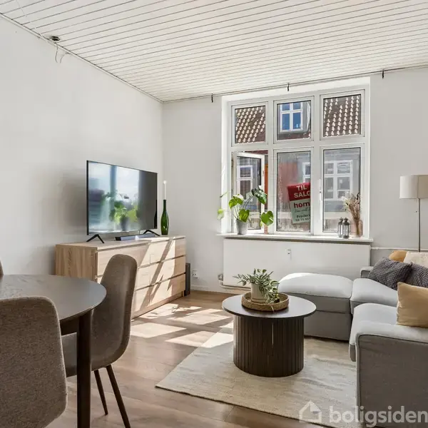 Et lyst køkken med hvide skabe og bordplader. Et lille bord med to stole er placeret nær vinduet, som viser udsigt over tage. En ovn og emhætte findes på højre side.