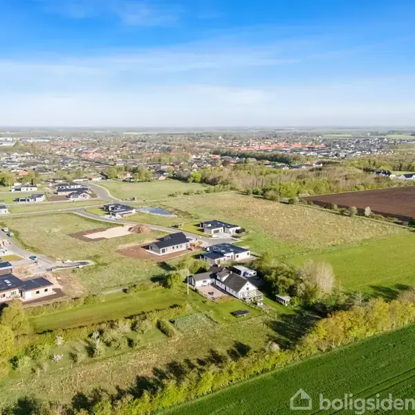 Landsbyhuse ligger spredt i et grønt landskab med marker og træer. I baggrunden ses en større by med flere huse og bygninger under en klar blå himmel.