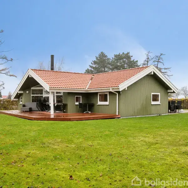 Grønt hus med rødt tag står stille omgivet af en veltrimmet græsplæne. En stor træterrasse med havemøbler ses foran huset. Skov og hække omkranser ejendommen.