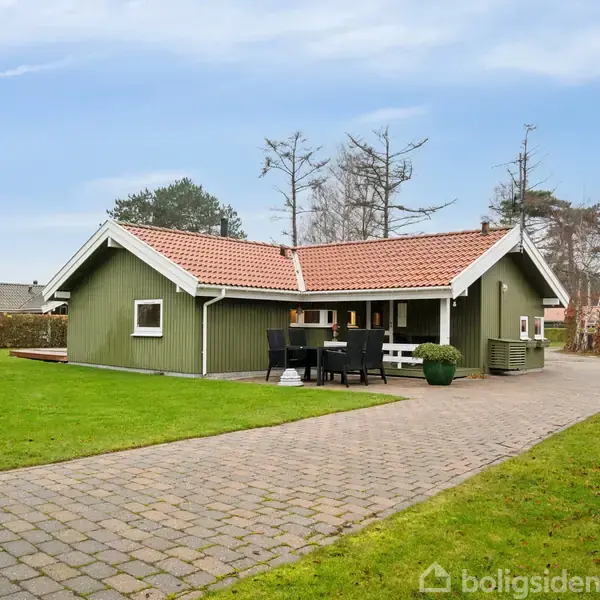 En grønmalet bungalow med rødt tag ligger i en velplejet have. På terrassen står et sort bord og stole. Baggrunden indeholder træer og en klar himmel.