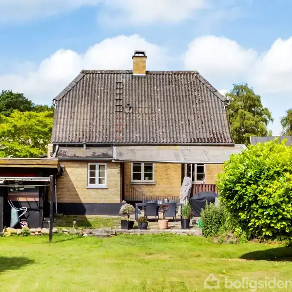 Hus med brun facade og skråt tag ligger i en have med en græsplæne. Der er en terrasse med planter. Omkranset af træer og hække.