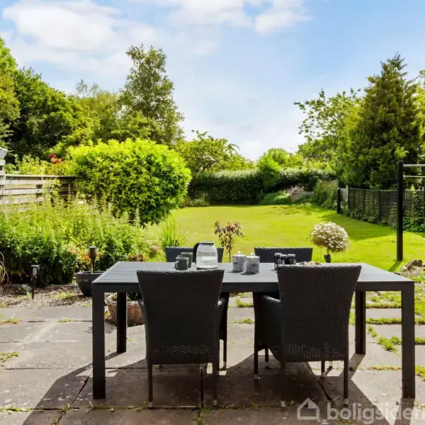 Et sort bord med fire stole står på en terrasse, omgivet af en grøn have med buske og hegn. Møblerne er placeret på fliser med udsigt til en stor græsplæne.