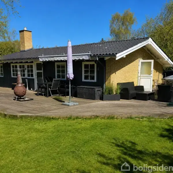 Et sommerhus med træterrasse har udemøbler og en stor parasol. Huset er omgivet af græsplæne og omgivet af træer under en klar blå himmel.