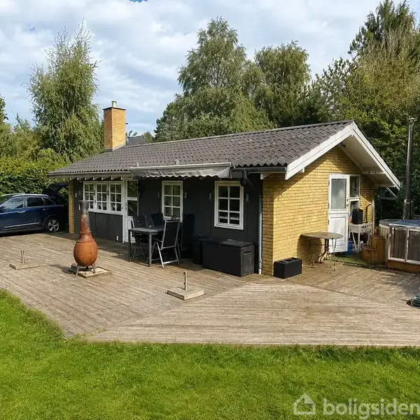Et gult murstenssommerhus med sort tag står på en stor træterrasse. Der er havemøbler, en bil parkeret til venstre og en græsplæne omkring, omkranset af træer.