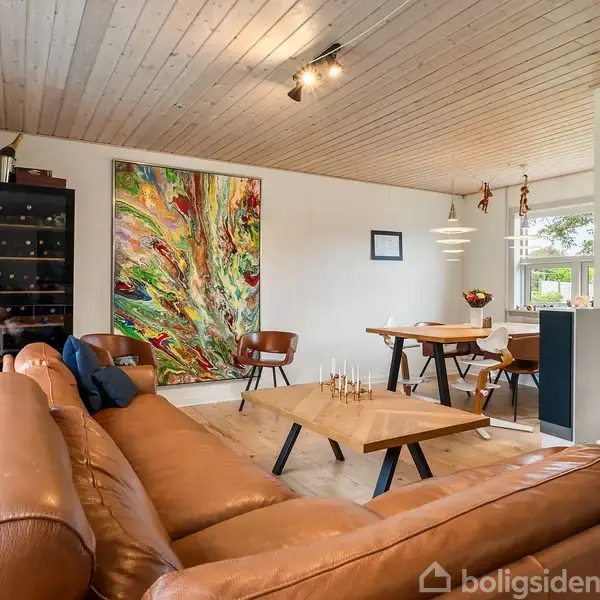 Lædersofa i brun står foran sofabord. Rummet har en stor, farverig vægkunst samt spisebord med stole. Loftet er træbeklædt, og der er store vinduer i rummet.